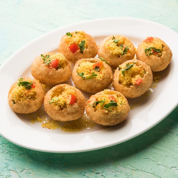 A white plate holds nine golden-brown pani puri, filled with yellow sev, red tomatoes, and green herbs.