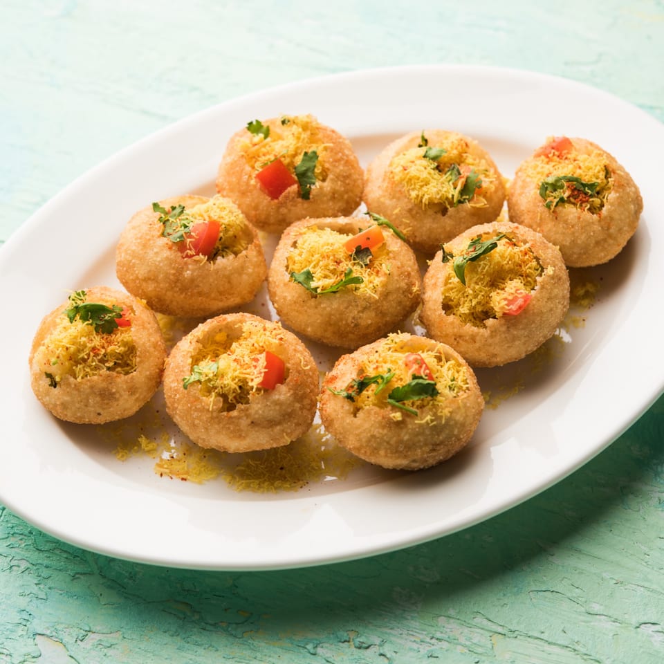 A white plate holds nine golden-brown pani puri, filled with yellow sev, red tomatoes, and green herbs.