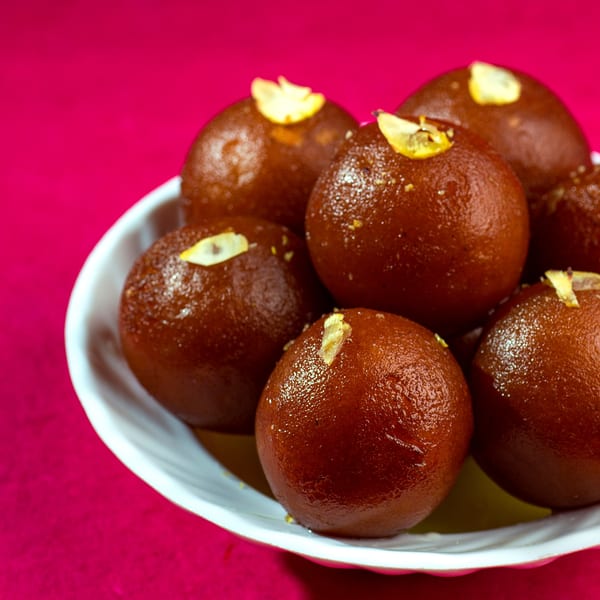 A white bowl filled with glistening Gulab Jamun, garnished with slivered almonds, on a vibrant pink background.